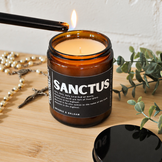 An amber glass jar candle labeled 'SANCTUS' with a black lid placed beside it on a wooden surface, while a black lighter burns the wick. Decorative beads, greenery, and a white brick wall are visible in the background.