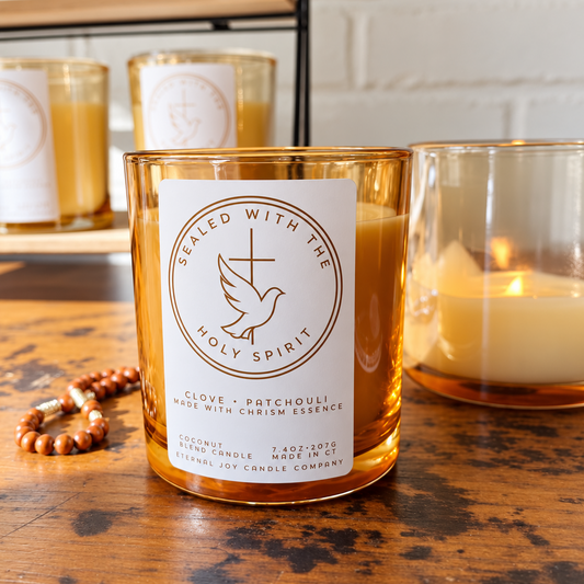 A clove and patchouli scented candle in an amber glass jar with a white paper label reading 'Sealed with the Holy Spirit,' placed on a wooden table with other candles and a beaded bracelet nearby.