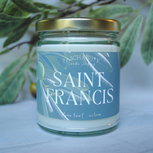 Candle jar labeled 'Saint Francis' with a gold lid on a white surface with green leaves in the background