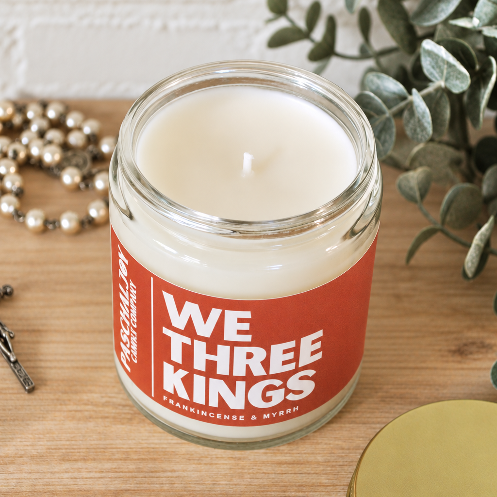A glass jar candle with cream-colored wax and a red label reading 'WE THREE KINGS' sits on a wooden surface with eucalyptus leaves, a gold lid, and a white brick wall in the background.