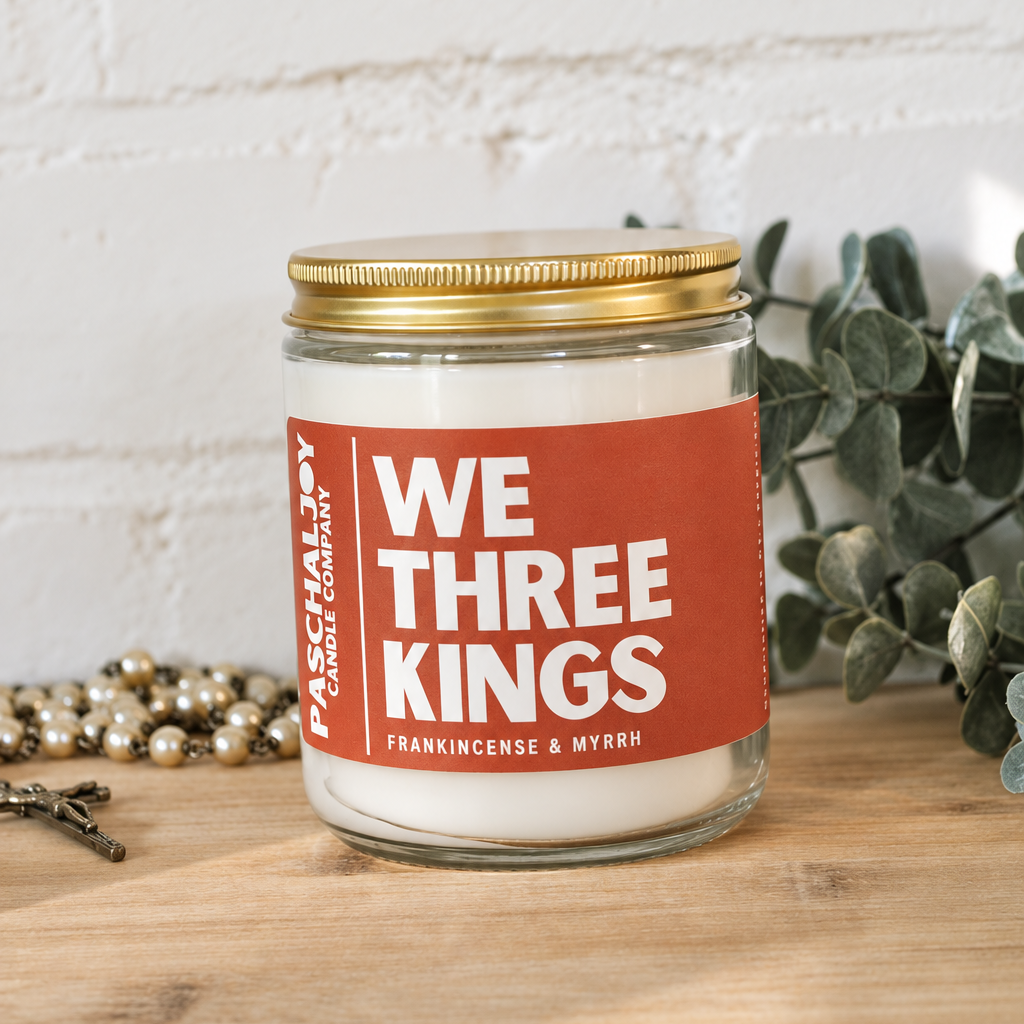 A glass jar candle with white wax and a gold metal lid sits on a wooden surface against a white brick wall. The candle has a terracotta red label with white text reading 'WE THREE KINGS' and smaller text 'FRANKINCENSE & MYRRH.