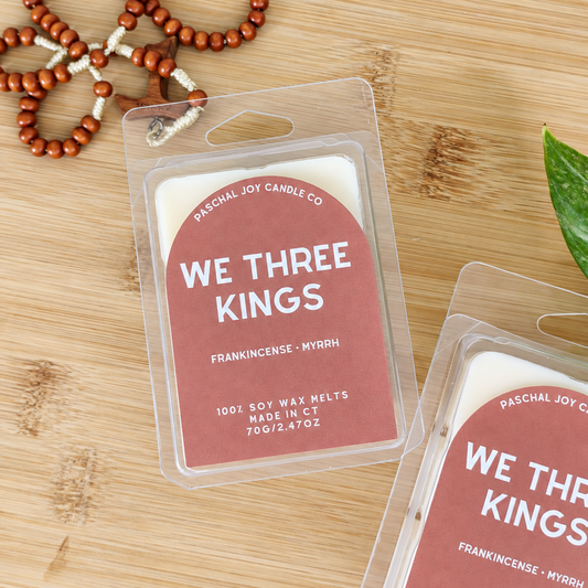 Two packaged soy wax melts from Paschal Joy Candle Co. labeled 'WE THREE KINGS' on a light wood surface, with a beaded bracelet and a green leaf nearby as styled props.