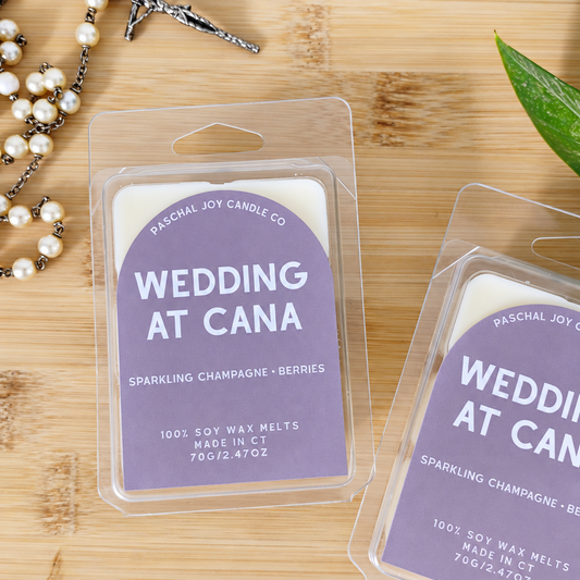 Two packaged soy wax melts from Paschal Joy Candle Co. labeled 'Wedding at Cana' in a lavender card with clear plastic clamshell packaging, arranged on a light wood surface with a pearl rosary and green leaves as props.