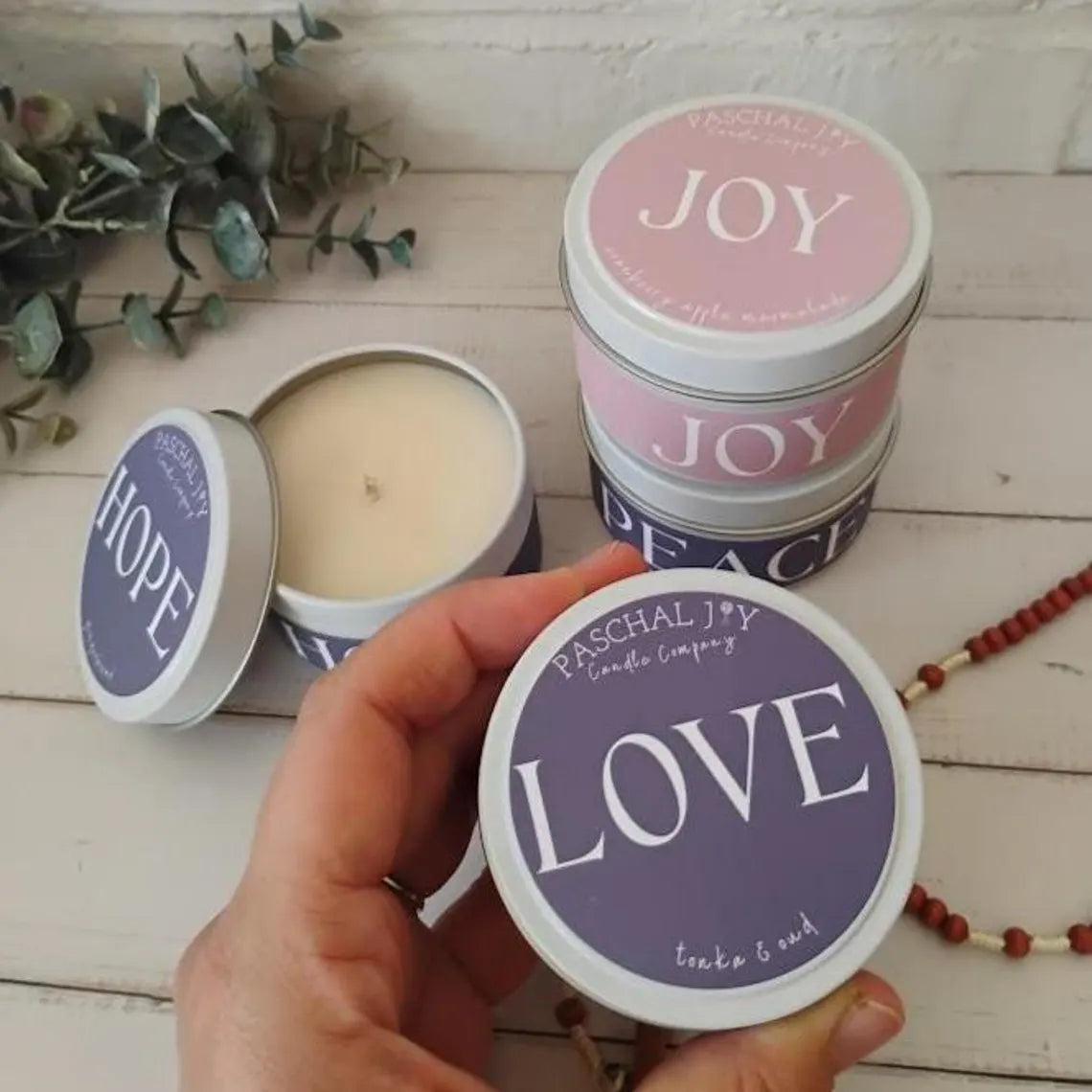 Candle tins with 'HOPE', 'JOY', and 'PEACE' on lids, held by a hand, with a 'LOVE' tin in focus.
