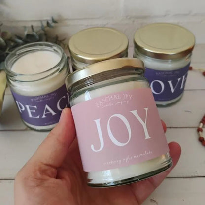 Advent Candle jars with labels 'Peace', 'Joy', and 'Love' held by a hand on a wooden surface.