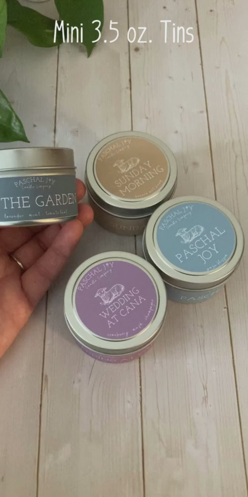 A hand is showing a scented tin candle called IN THE GARDEN. Three candle tins are in the background. Light grey wood background.